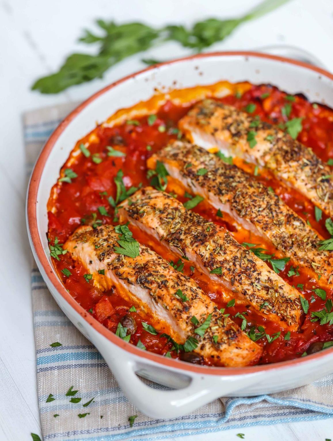 Mediterranean baked salmon fillets in a rich tomato and caper sauce in a white baking dish, garnished with chopped parsley